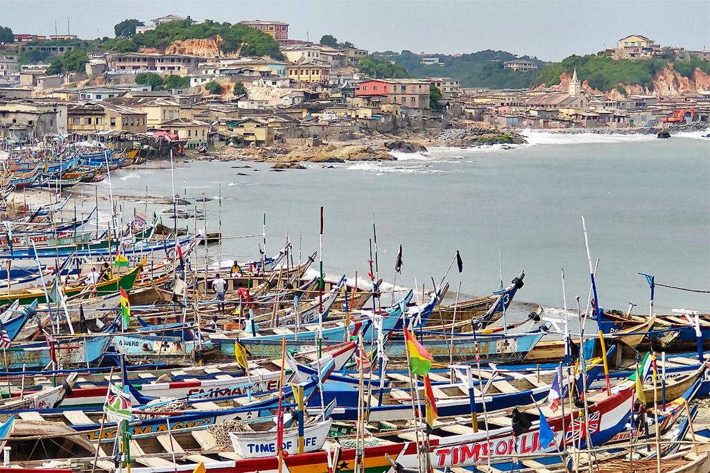 Blick vom Cape Coast Castle auf zahlreiche bunt bemalte Fischerboote am Ufer und die Häuser entlang der Bucht.