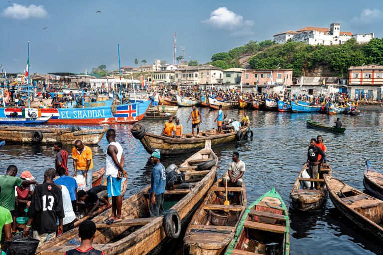 Fischerboote und geschäftiges Treiben am Fischereihafen von Elmina in Ghana, mit Elmina Castle im Hintergrund.